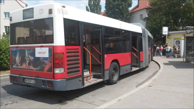 Hochflur-Gelenkautobus mit geöffneten Türen in einer Station. In der hintersten Tür sind die Stufen zu sehen. Im Hintergrund steigen Menschen in den vorederen Teil des Busses ein.