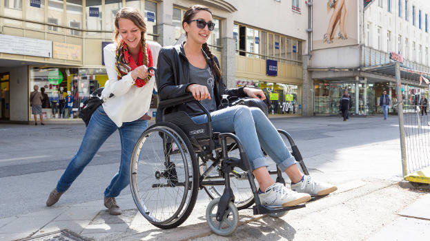 Eine junge Frau im leichten Rollstuhl wird von einer anderen jungen Frau über die Gehsteigkante geschoben.
