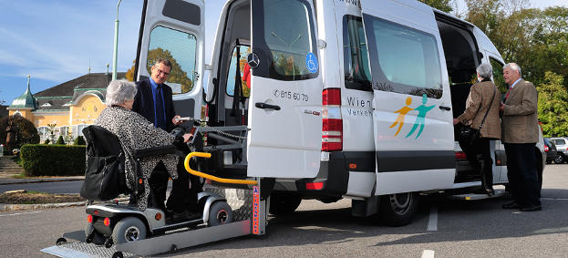 Ein Van des Sonderfahrtendienstes der WLV mit dem Heck zum Betrachter. Im Heck wird eine Dame im Rollstuhl mit einer Hebeplattform in den Wagen gehoben. Ein freundlich lächelnder Herr steht daneben. Eine Dame steigt bei der Mitteltüre ein, während ihr ein anderer freundlicher Herr den Ellbogen stützt.